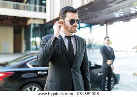 Handsome Bodyguard Listening To Earpiece With Car Parked On Street In City