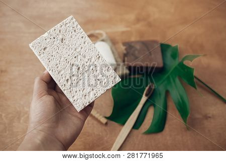 Hand Holding Natural Eco Sponge On Background Of Bamboo Toothbrush, Soap, Solid Shampoo, Crystal Deo
