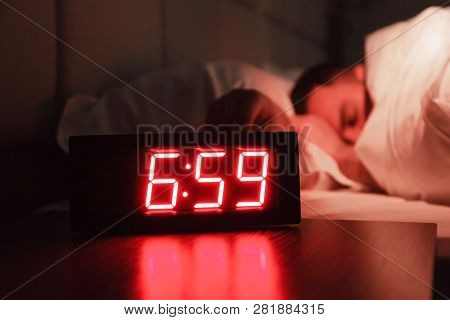 Rectangular Alarm Clock On The Bedside Table With Red Numbers, Sleeping Man In Bed In Dark Room. Con