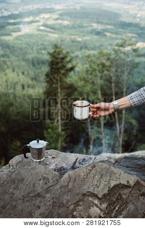 Girls Hand Holds A Hot Cup Of Coffee Near The Campfire On Background Of Nature. Concept Adventure Ac