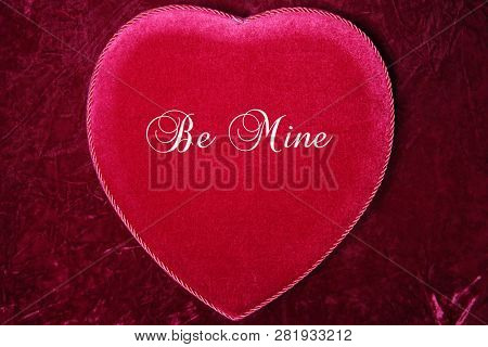 Valentines Day. Red Velvet Heart Shaped Valentines Day Chocolates Box.
On Burgundy Red Velvet background.  Text reads, Be Mine

