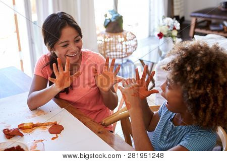Two young teen and pre-teen girlfriends sitting at a table at home using modelling clay, showing each other their dirty hands, elevated view