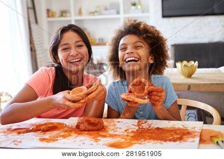 Two girlfriends having fun playing with modelling clay at home, laughing and showing their creations to camera, front view, close up