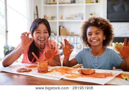 Two girlfriends having fun playing with modelling clay at home, smiling and showing dirty hands to camera, front view, close up