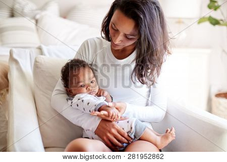 Young adult mother sitting in an armchair in her bedroom, holding her three month old baby son in her arms and looking down at him, close up