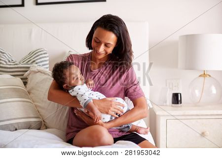Millennial mixed race mother  sitting on her bed holding her three month old baby son in her arms