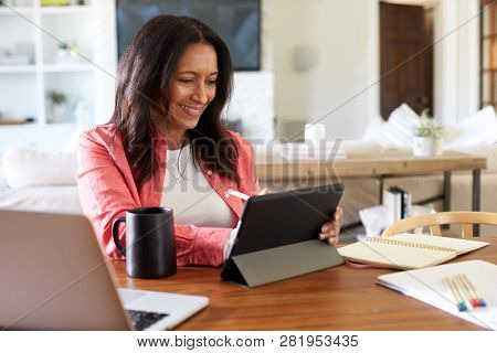 Smiling middle aged woman sitting at the table in her dining room drawing on a tablet computer with a stylus, close up