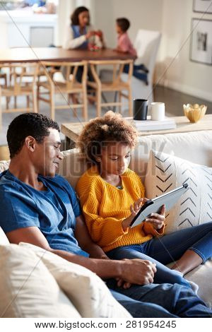 Pre-teen girl sitting on sofa in the living room using tablet computer with her father, mother and toddler sitting at a table in the background, selective focus, vertical