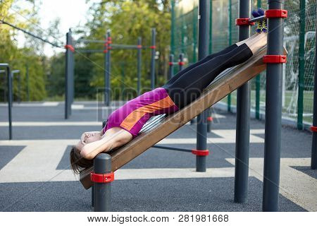 Young Caucasian Woman Workouts On The Park Sports Ground. The Girl Does Abdominal Excersise, In Blac