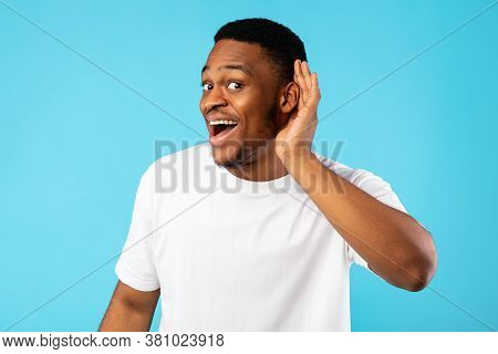 Positive African Man Listening Holding Hand Near Ear Posing On Blue Studio Background. Huh, I Hear Y