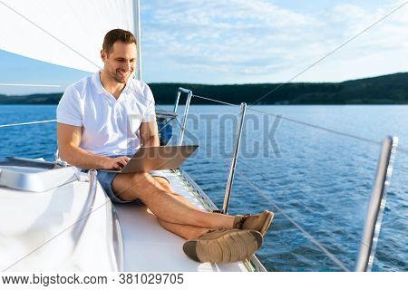 Happy Man On Yacht Sitting With Laptop Working Living On Sailboat During Summer. Liveaboard Tour. Em
