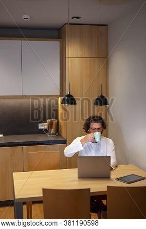 Young Man Sitting At Dinning Room Table, Wearing Pajamas, Drinking Coffee While Working Overtime, Us