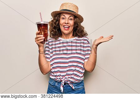 Middle age brunette woman on summer hat drinking fresh cola refeshment over isolated background celebrating achievement with happy smile and winner expression with raised hand