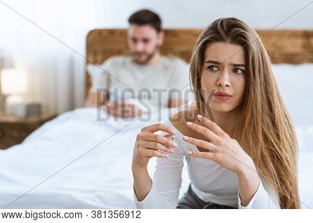Family Problems And Divorce. Sad Wife Takes Off Her Wedding Ring Close Up, Husband Typing On Phone I