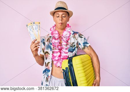 Young hispanic boy wearing summer style holding cabin suitcase and boarding tickets making fish face with mouth and squinting eyes, crazy and comical. 