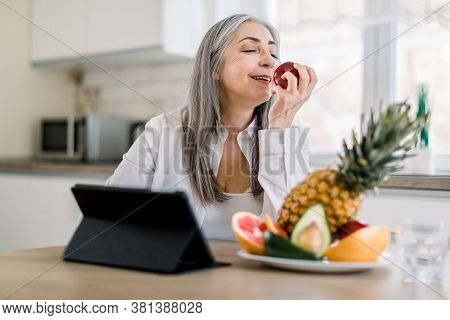 Beautiful Elderly Female Pensioner With Long Gray Hair Sitting At The Kitchen Table At Home And Enjo