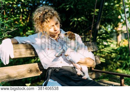 Graceful Blond Haired Woman Gardener In White Shirt Sitting On The Bench At Yard With Puppy Dog Rela