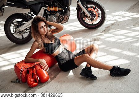 Attractive Sportswoman Posing In A Light Studio Lying On The Floor With A Large Orange Punching Bag.