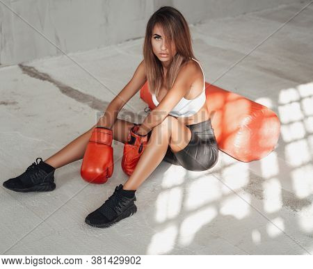 Young And Beautiful Sportswoman Posing In A Light Studio Sitting On The Floor With A Large Orange Pu