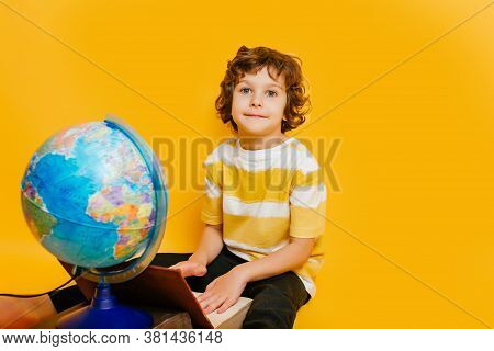 Curly Boy In Glasses And Stripes Tshirt And Sits On A Stack Of Books, Near To The Globus On Yellow B