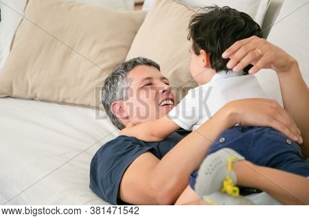 Cheerful Father Lying On Sofa With Adorable Little Boy. Happy Attractive Caucasian Dad Hugging Or Em