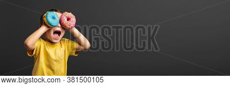 Happy cute boy is having fun played with donuts on black background wall. Bright photo of a boy. Colored donuts