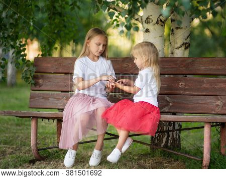 Two Small Sisters Are Sitting On Wooden Bench And Lplaying In The Park