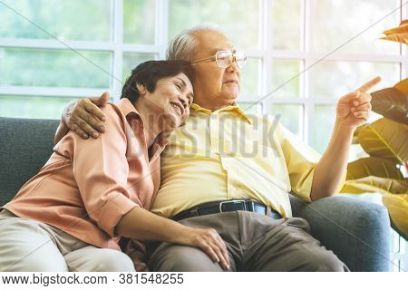 Happy Senior Couple Is Sitting Hugging Each Other On Sofa At Home Looking Out The Windows Peacefully