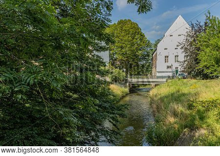 Jeker River With Crystal Clear Water Between Green Leafy Trees With A Vehicular Bridge And A White H
