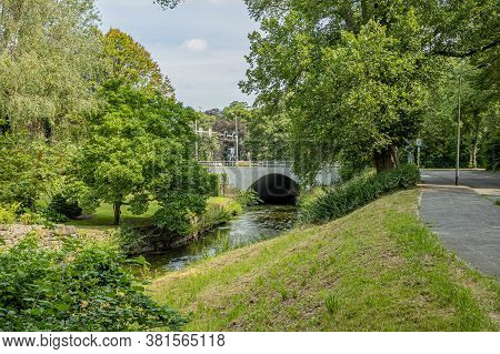 Jeker River Between Green Grass, Trees And Abundant Vegetation, A Vehicular Bridge With A Metal Fenc