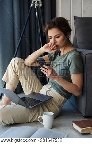 Attractive young woman sitting at the couch on a floor of a living room, using laptop computer, using ombile phone
