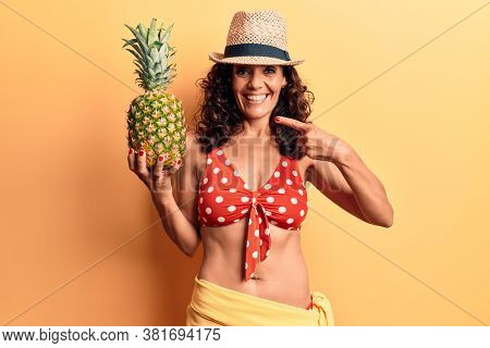 Middle age beautiful woman wearing bikini and summer hat holding pineappel smiling happy pointing with hand and finger 