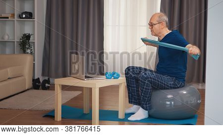 Old Man On Balance Ball Training With Resistance Band Watching Online Fitness Program. Old Person Pe
