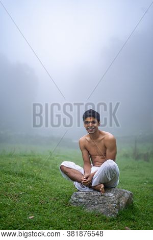 Young Man Wearing A Dhoti, Sitting On A Rock, Open In The Green Field In A Foggy Morning.