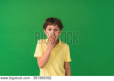 Little Boy Standing On A Green Isolated Background Covers His Mouth With His Hands In Shock For The 