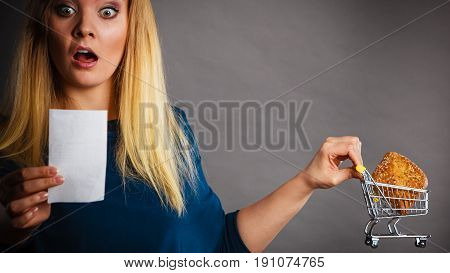 Shocked woman holding shopping basket with bread looking at bill receipt being scared of huge prices