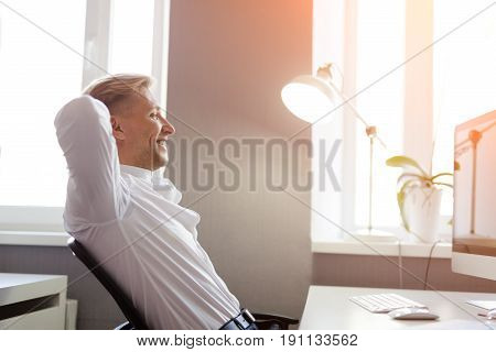 Side view of young content man sitting at desktop with computer and relaxing.