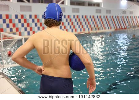 Young man in a cap with a ball for water polo looks at the game in the pool, view from the back