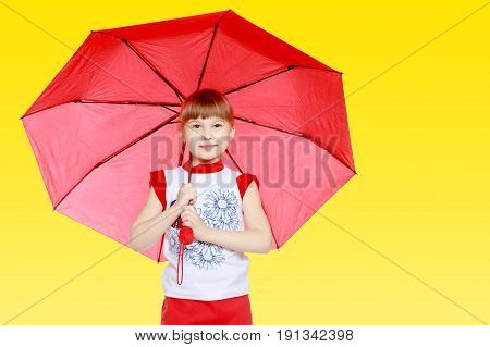A nice little blonde girl with a short bangs. In a red skirt and a white T-shirt with a pattern. The girl hid under the red umbrella.