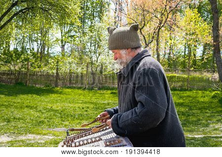 Kyiv Ukraine - April 23 2017: An elderly Ukrainian man a peasant with a long mustache and beard plays an ancient musical instrument of cymbals. Spring landscape. Old Street Musician. Museum Pyrohiv
