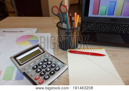 Close up detail view of a white work desk with a laptop computer and a calculator with numbers on the screen. Working analyzing and doing calculations