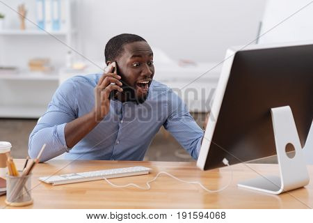 Great news. Happy cheerful Afro american man looking at the computer screen and showing his happiness while speaking on the phone