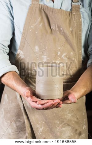 pottery, stoneware, ceramics art concept - young man in a dirty apron stand at workshop, master hands holds cup of unfired clay, male examines product before further processing, front view, vertical