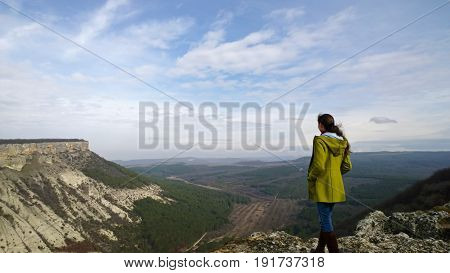 Brunette girl in a bright green coat standing on top of the old city Chufut-Kale. Hands in pockets, hair up in a ponytail. The view from the back