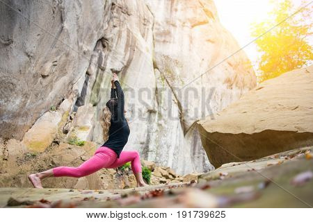 Flexible Fit Female Is Practicing Yoga And Doing Asana Virabhadrasana 1 Outdoors. Huge Rocks On The