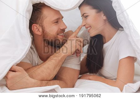 Got your nose. Adorable young couple lying underneath the blanket and having a pillow talk while a man touching his wife nose.