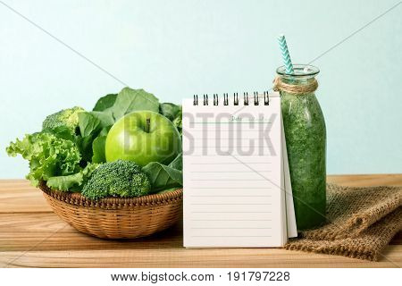 blank note pad and the Healthy fresh green smoothie juice in glass bottle on wooden table with green apple and vegetables basket for healthy detox and diet habits concept