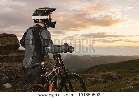 Ryder in full protective equipment on the mtb of two pendant bicycles rides along the green grass towards the sunset in the rays of the sunset sun against the backdrop of a landscape of rocky terrain under the setting sun and clouds.