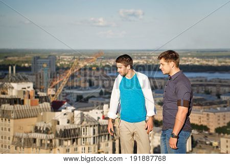 Two roofers on the roof of a high-rise building admire the view of the city. Courage and adrenaline. The men are looking down.