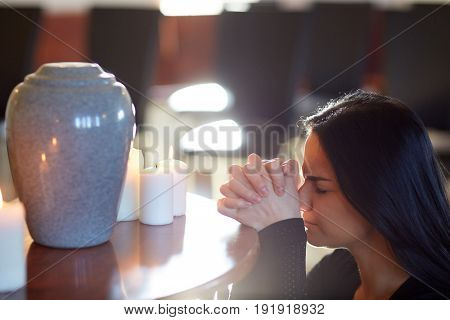 people and mourning concept - sad woman with cinerary urn and candles praying at funeral in church